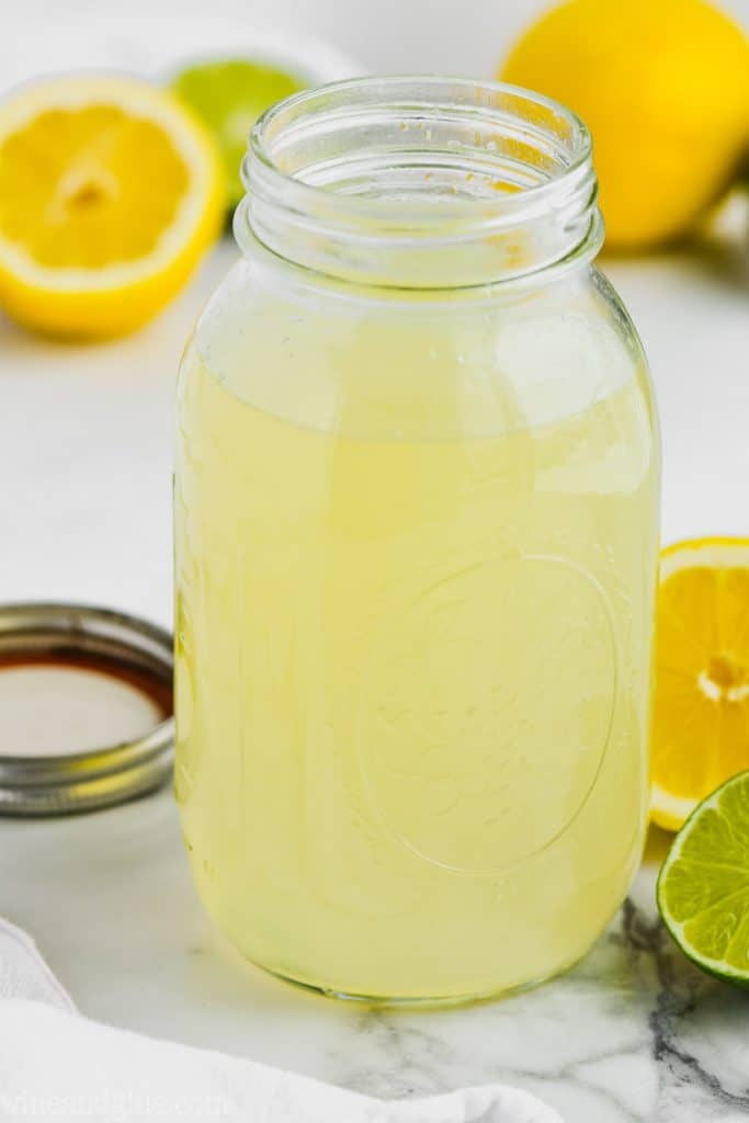 clear mason jar full of sweet and sour mix with limes and lemons around it on marble counter top