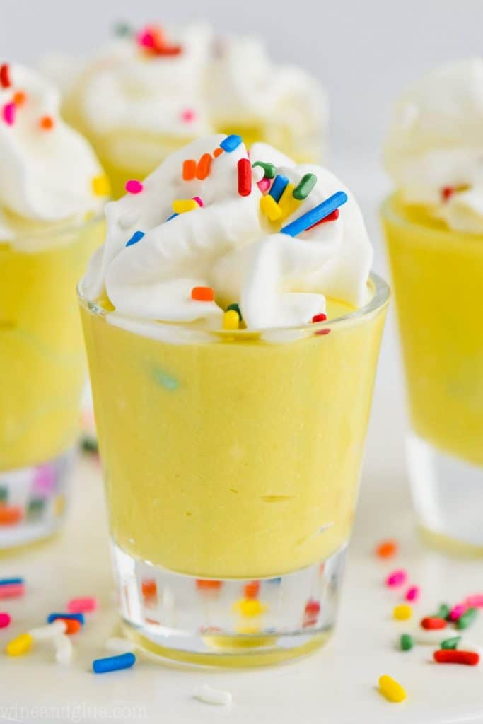 straight on view of a shot glass filled with birthday cake pudding shots and topped with whipped cream and rainbow sprinkles, other shot glasses in the background