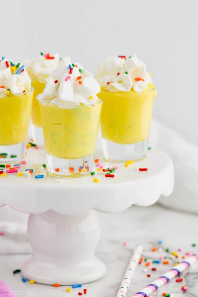 four shot glasses filled with yellow birthday cake pudding shots and topped with whipped cream and rainbow sprinkles on a small cake stand