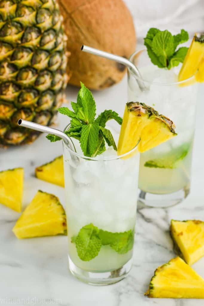 two high ball glasses filled with pineapple mojitos, garnished with pineapple wedges, and fresh mint with a pineapple and a coconut in the background