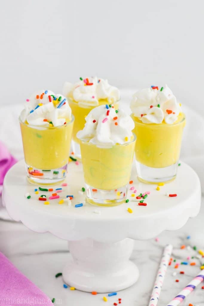 four shot glasses filled with yellow birthday cake pudding shots and topped with whipped cream and rainbow sprinkles on a small cake stand