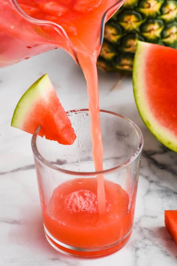 watermelon rum punch being poured into a small tumbler glass that is garnished with a watermelon wedge