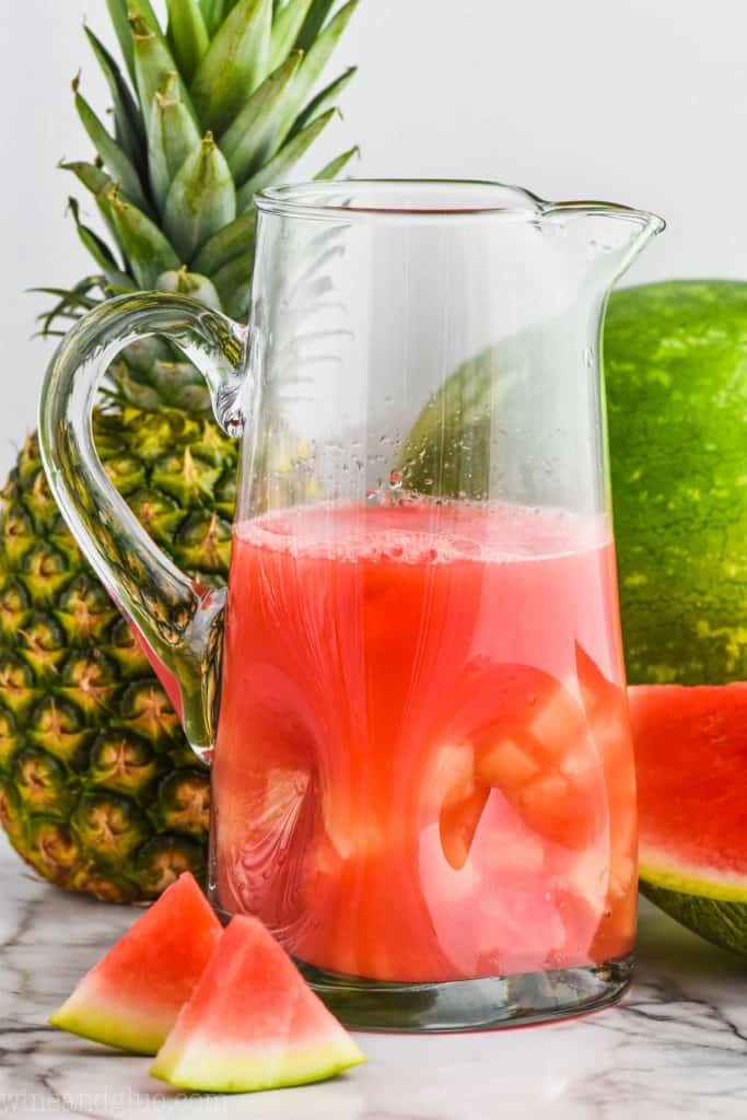 a tall pitcher of watermelon rum punch with a pineapple and a watermelon in the background