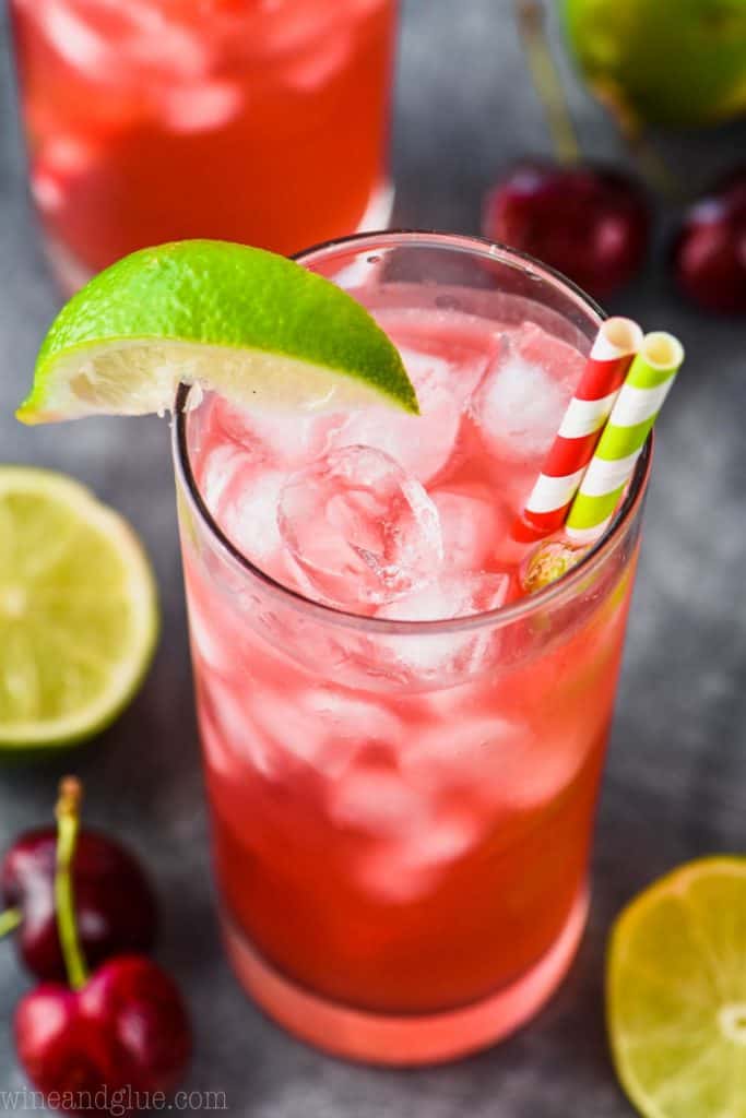 overhead view of a high ball glass with ice, cherry lime vodka tonic, a striped red straw, a striped green straw and a lime wedge