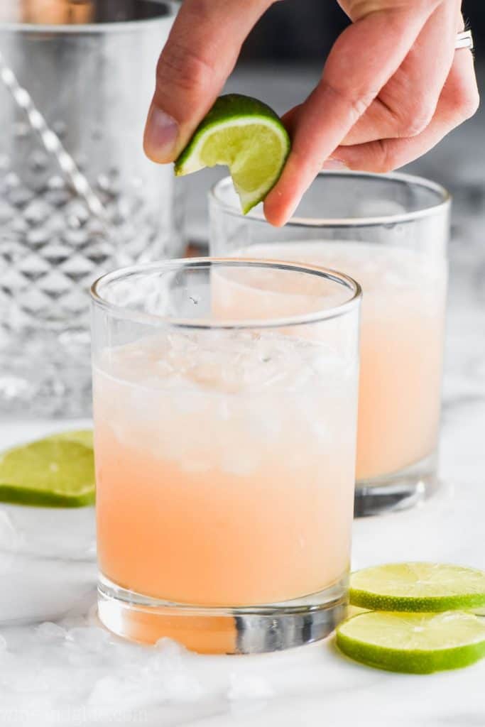 fingers squeezing a lime wedge into a tumbler half filled with ice and a paloma cocktail