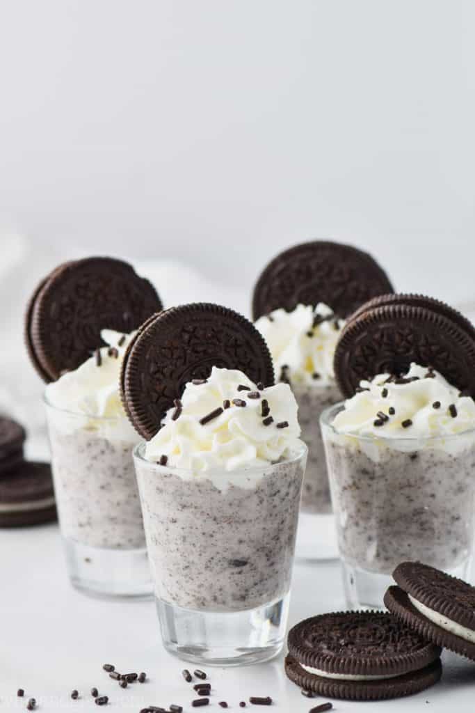 four shot glasses filled with cookies and cream pudding shots and garnished with oreo cookies, whipped cream and sprinkles.