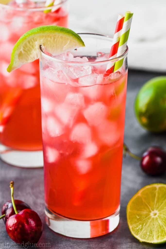 high ball glass full of bright red cherry limeade vodka tonic recipe, garnished with a red striped straw, a green striped straw and a lime wedge
