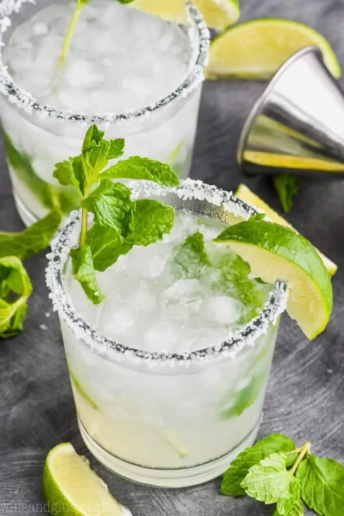 two tumbler glasses filled with a combination of mojito and margarita, rimmed with salt and garnished with a lime and mint