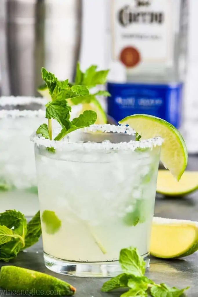 straight on view of a small tumbler of mojito margarita with a lime wedge and garnished with mint, tequila in the background