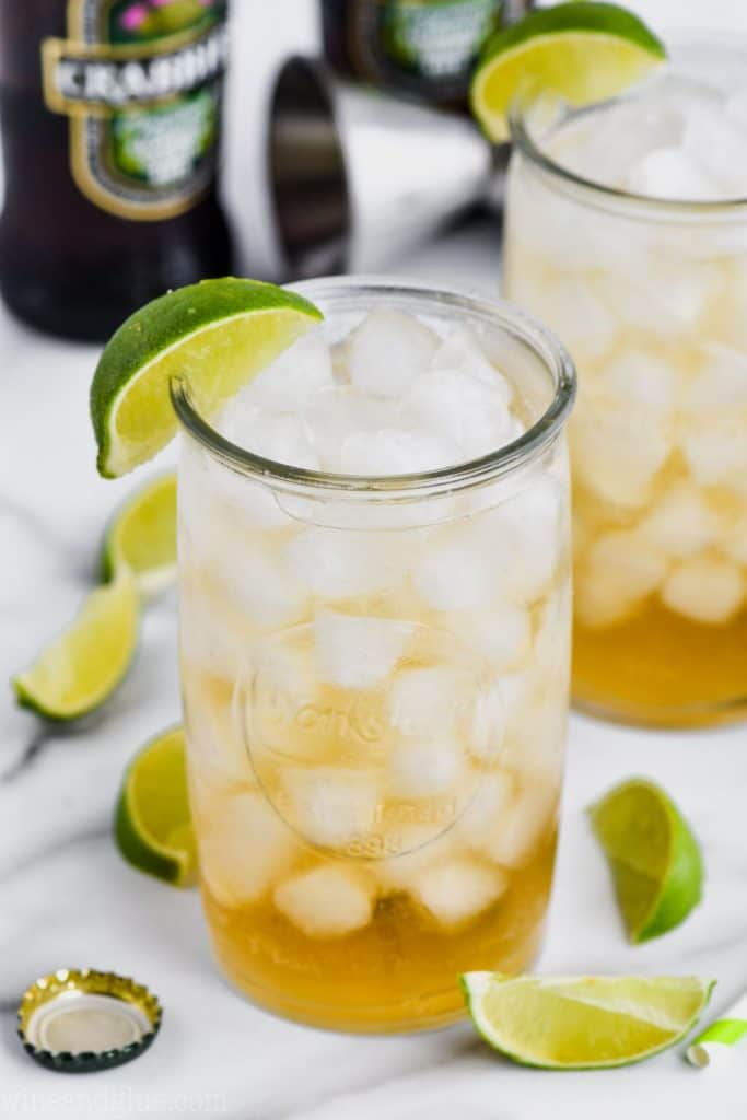 overhead angled view of a tall glass (another in the background) filled with ice and dark and stormy drink recipe, lime wedges near the base of the glass and ginger beer bottle in the background