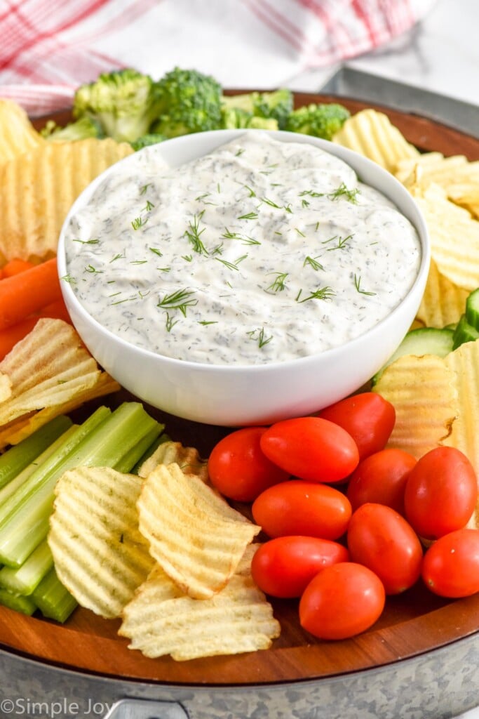 A white bowl full of dill dip on a platter with vegetables and chips