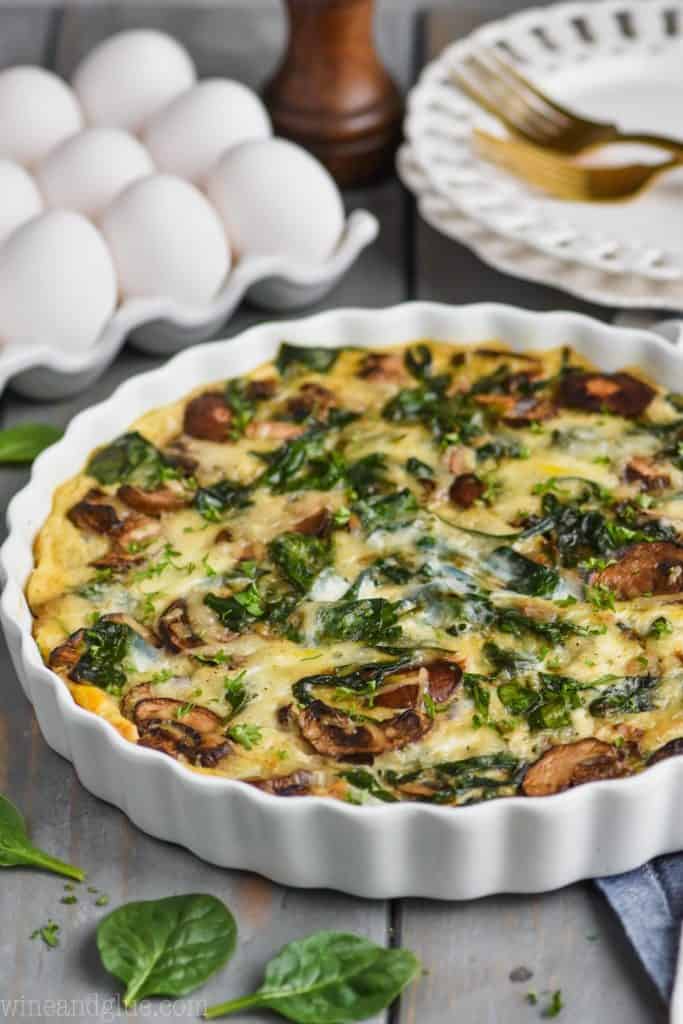 a spinach mushroom quiche in a white quiche dish with eggs in the background