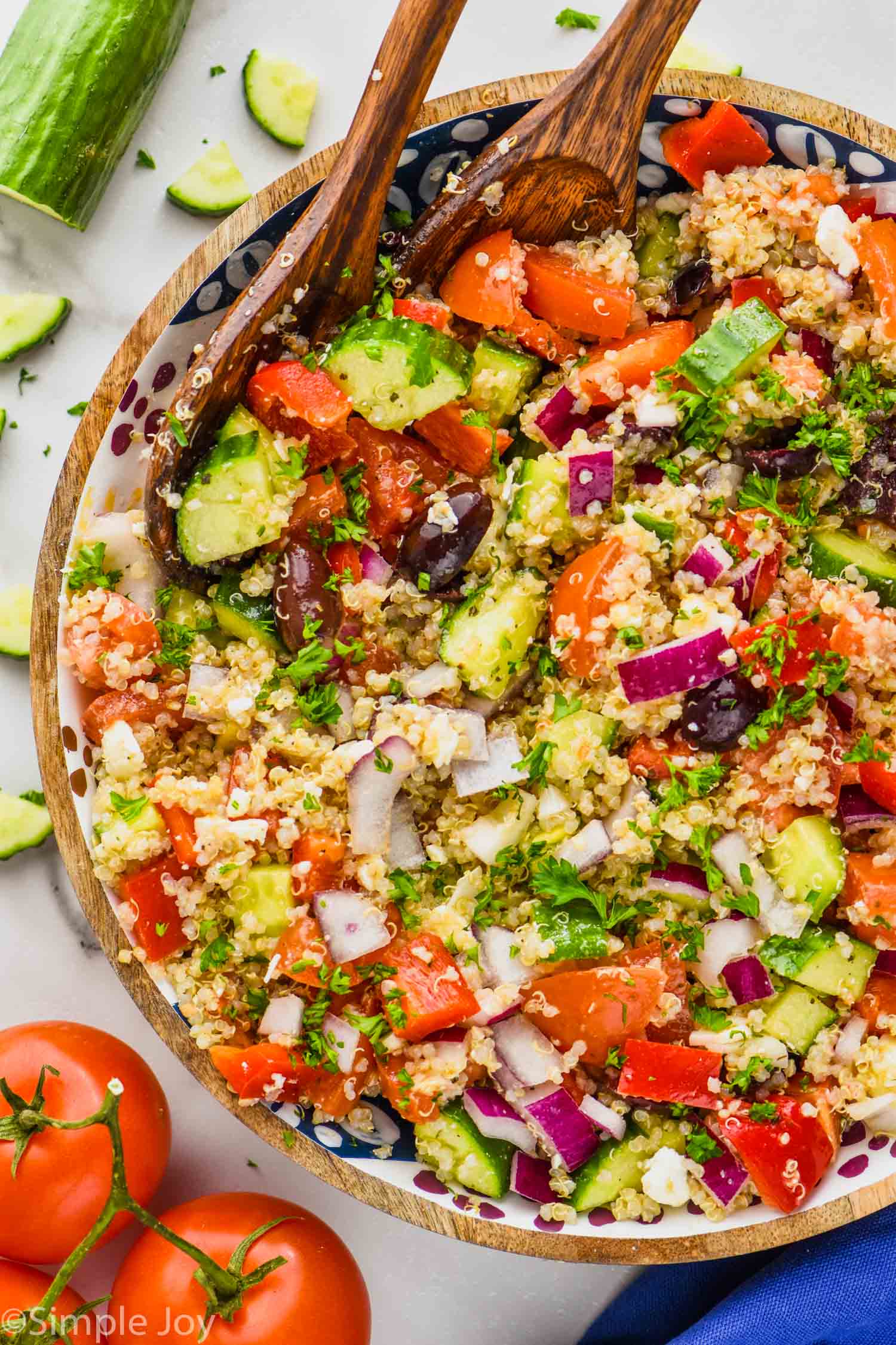 overhead view of a bowl of greek quinoa salad garnished with parsley, with cucumbers and tomatoes off to the side of the bowl