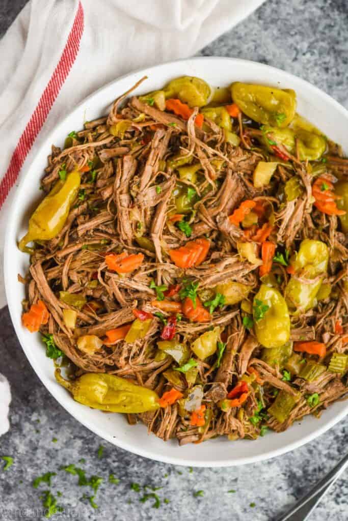 overhead view of italian beef recipe in a white bowl on a granite surface with a cloth napkin next to it