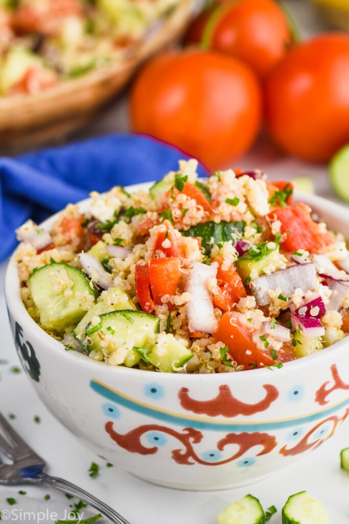 a small bowl of mediterranean quinoa salad garnished with parsley with tomatoes in the background