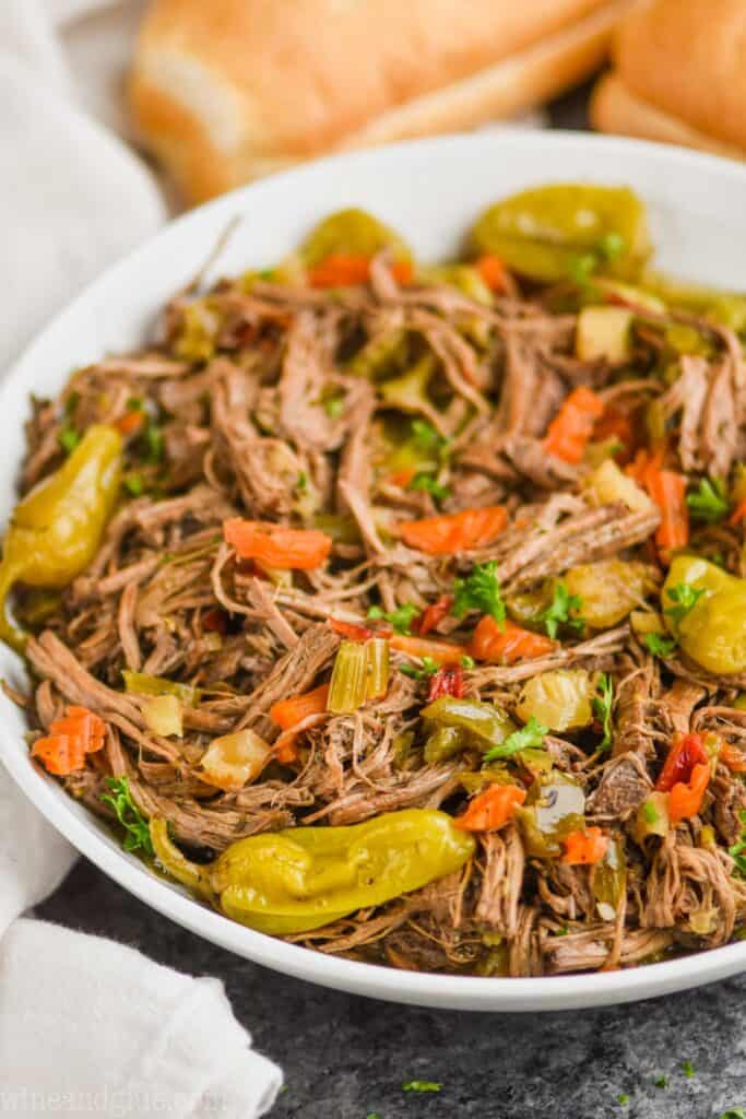 side angle view of slow cooker italian beef in a white serving bowl with hoagie rolls in the background