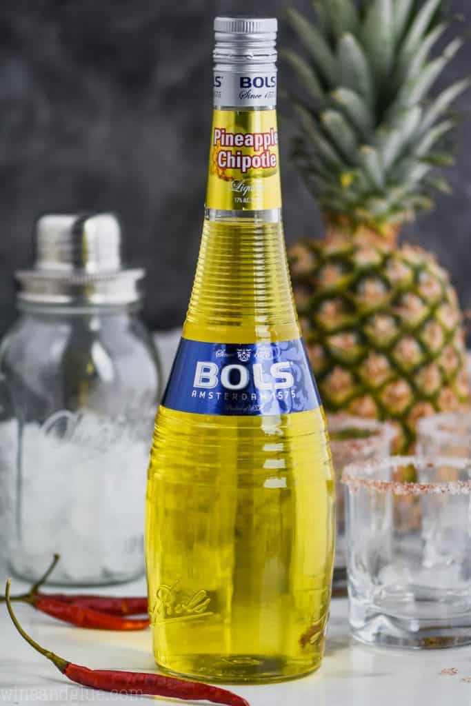 bottle of bols pineapple chipotle liqueur surrounded by glasses, a cocktail shaker, chiles, and a fresh pineapple