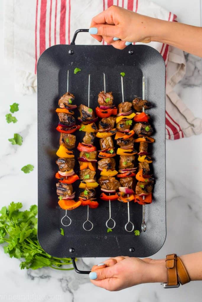 overhead view of hands holding a black tray with five steak fajita kabobs