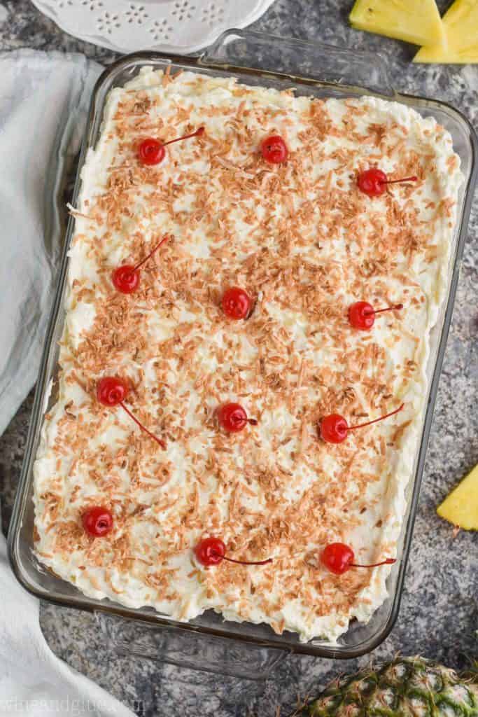 overhead view of 9 by 13 inch pan of poke cake topped with frosting toasted coconut and 12 cherries