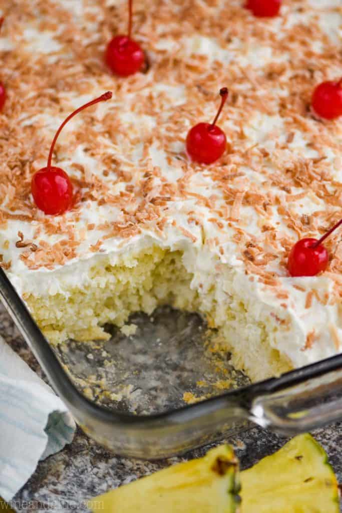 a sheet cake that has been made into a pina colada poke cake with a piece missing, frosted with toasted coconut on top and maraschino cherries