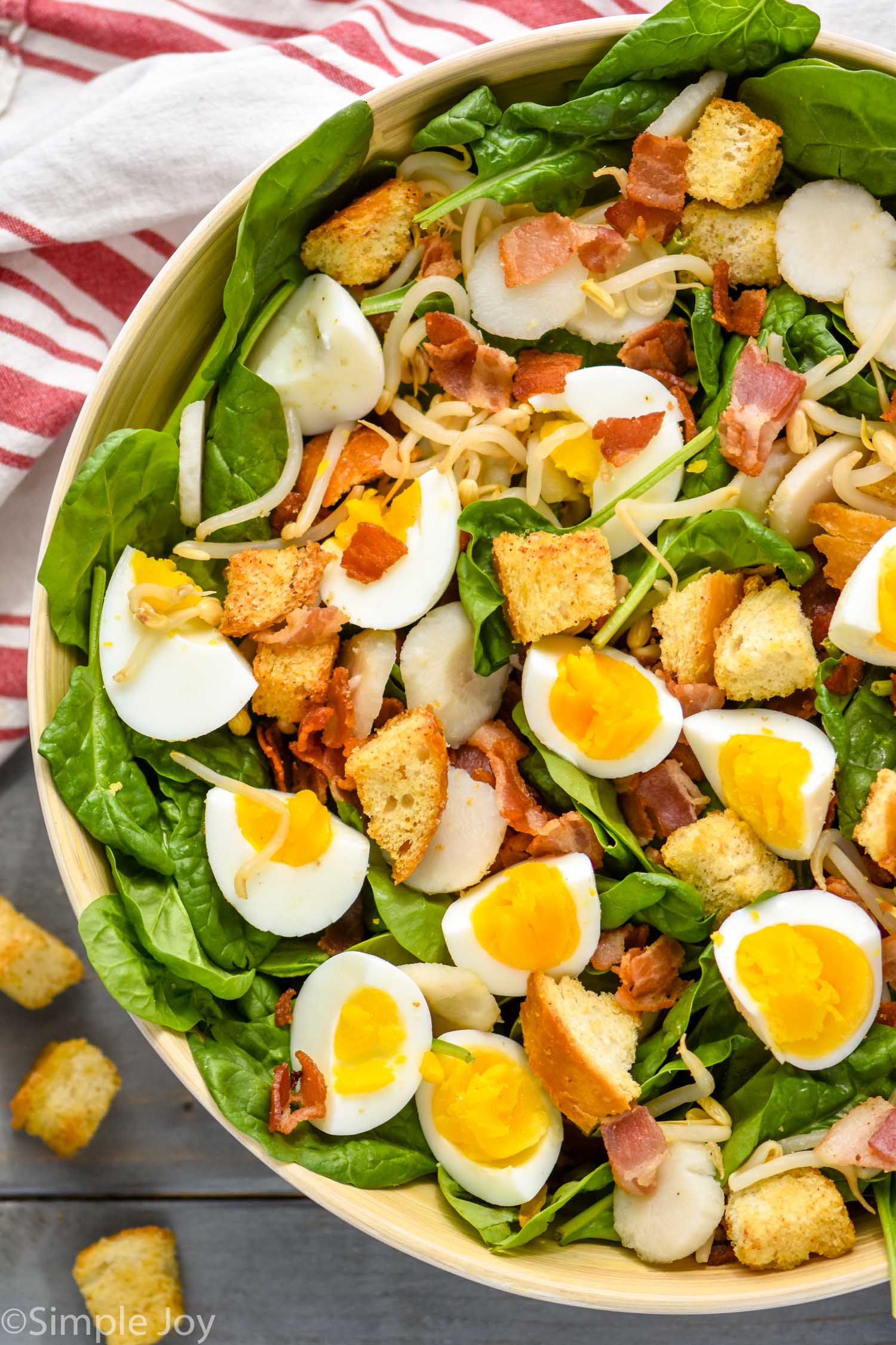 overhead view of spinach bacon salad with hard boiled eggs, croutons, water chestnuts in a white bowl