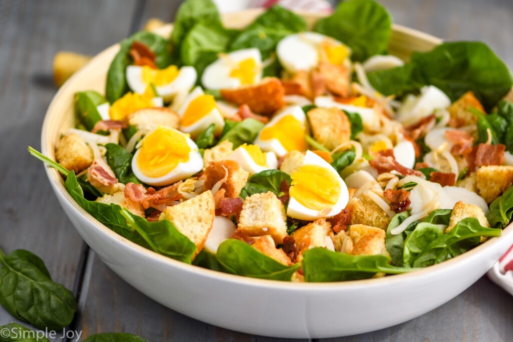 white bowl filled with spinach salad with bacon, hard boiled eggs, and croutons on a blue wood board