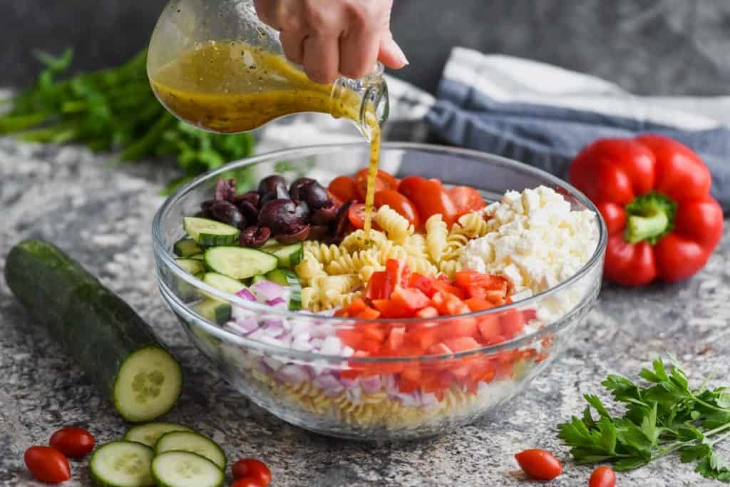 hand pouring greek salad dressing over a greek pasta salad in a clear bowl