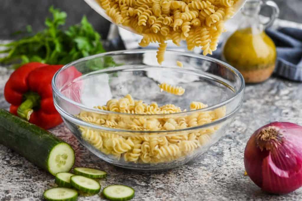 cooked noodles being poured into a bowl for greek pasta salad