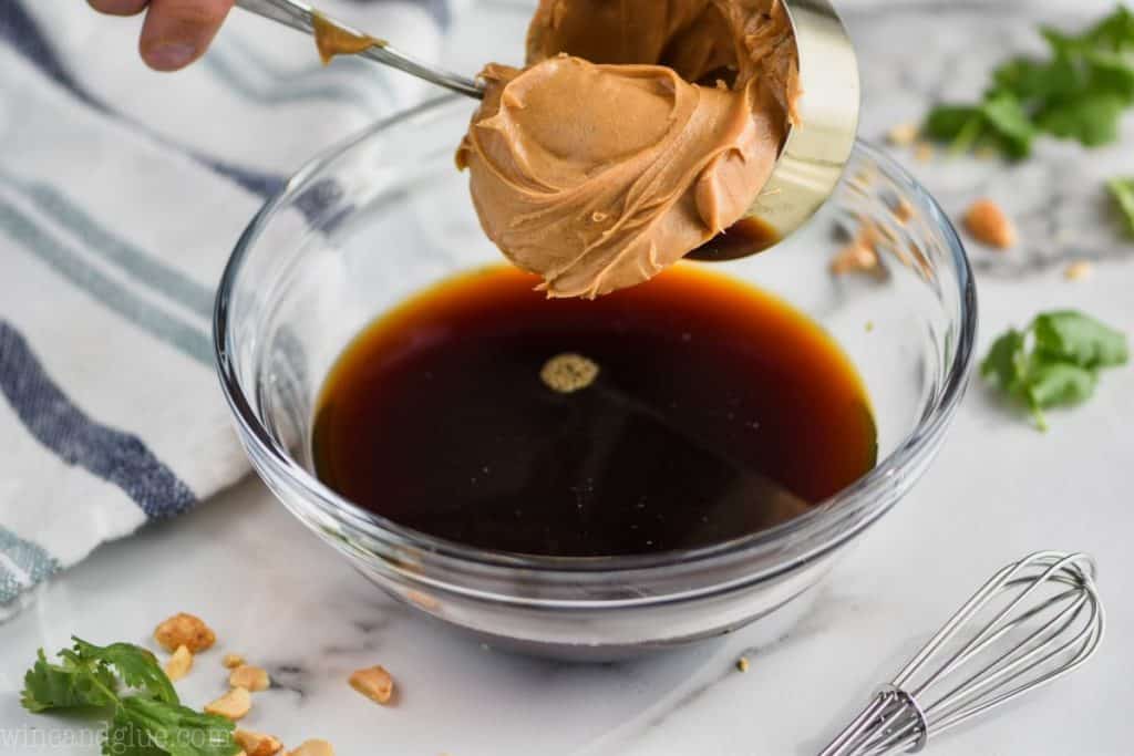 creamy peanut butter being put into a bowl filled with apple cider vinegar and soy sauce