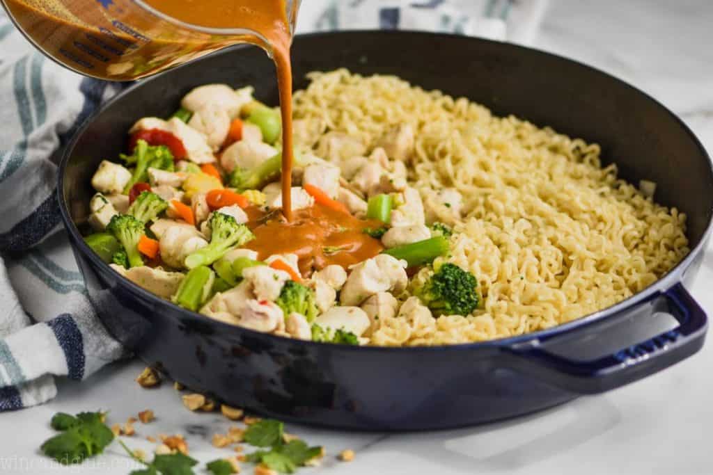 pouring peanut sauce into a skillet that contains vegetables and noodles