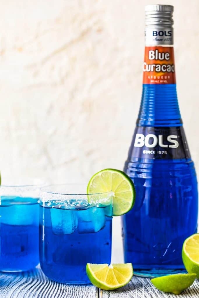 two glasses filled with blue margaritas, rimmed with salt, ganrished with a lime and with a bottle of bols blue curaçao next it