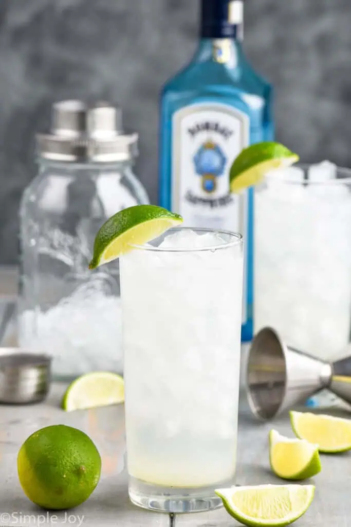 pulled back view of two glasses full of gin rickey recipe with shaker and bottle of Bombay sapphire gin in the background