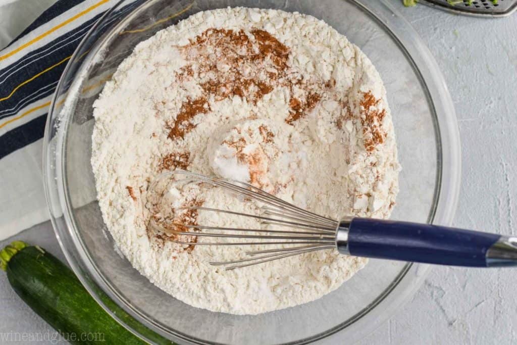 flour, baking soda, salt, and cinnamon being whisked together in a clear glass bowl to make zucchini muffins