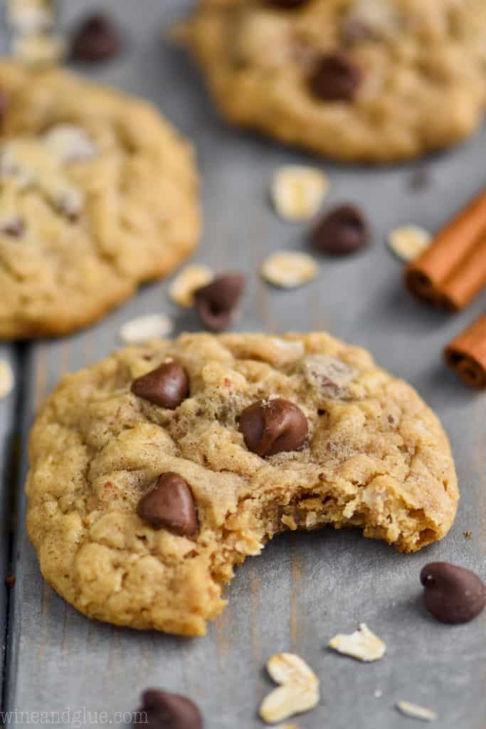 oatmeal chocolate chip cookie sitting on a blue wood board with chocolate chips and oats around it and two cinnamon sticks in the background