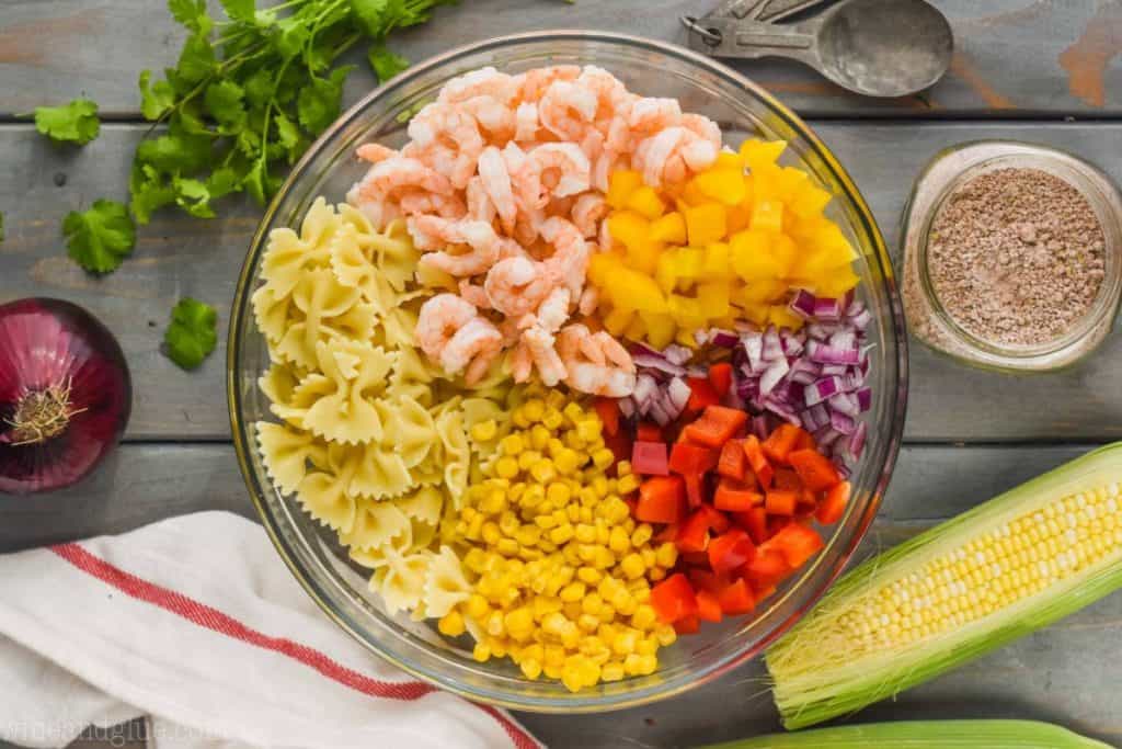 overhead view of a clear bowl filled with bow tie pasta, corn, red bell pepper pieces, red onion, yellow bell peppers, and shrimp broken down by ingredient 