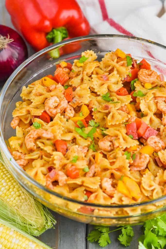 a clear bowl filled with shrimp fajita pasta salad, two ears of corn in front of it, some cilantro and a red bell pepper and a red onion behind it