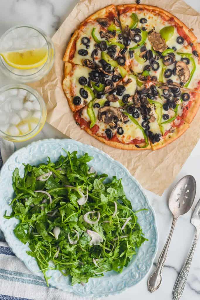 overhead view of arugula salad on an oval plate next to a 10 inch cauliflower crusted pizza topped with peppers, mushrooms, and olives