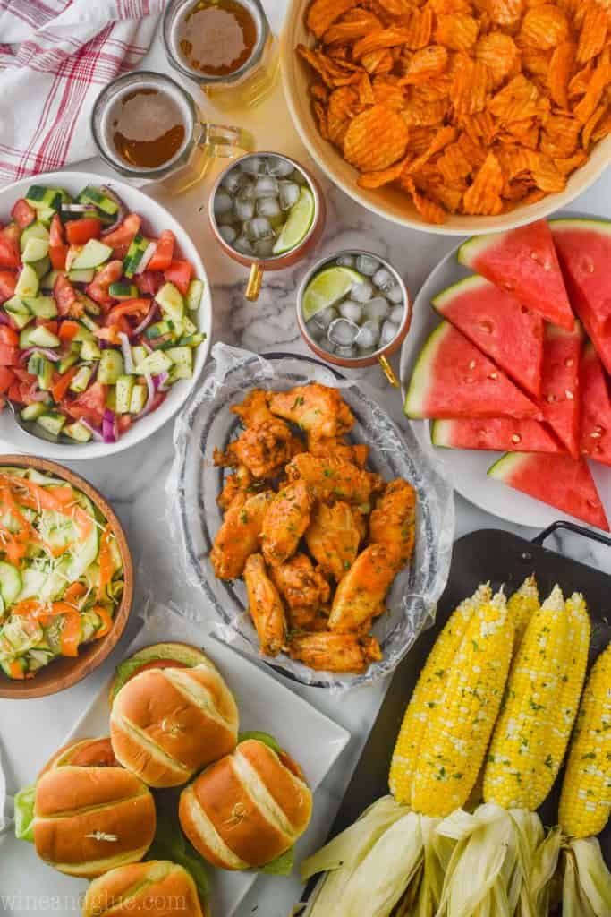 overhead view of a table filled with food for a party: crock pot chicken wings in the middle, corn that was grilled, fresh watermelon, potato chips, salads, and hamburgers