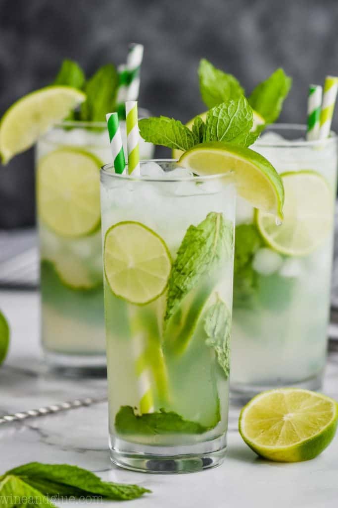 three high ball glasses on a white surface against a gray background filled with lime slices, a mojito recipe, fresh mint leaves, and garnished with green and white stripped straws, fresh mint and a lime wedge