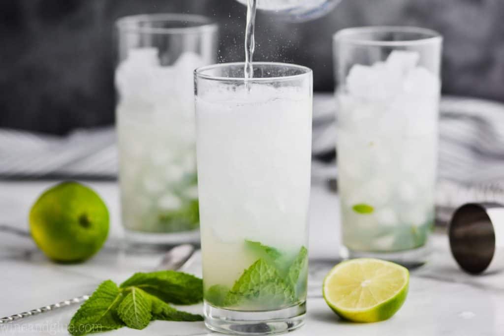 three glasses filled with ice, with mint and lime infront of the first glass and club soda being poured into it to make a mojito cocktail
