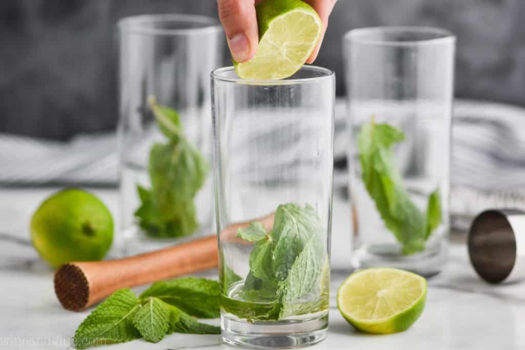 three high ball glasses with mint in them and a lime being squeezed into the first one to make a mojito drink