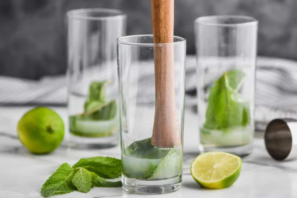 three high ball glasses with mint and lime juice and simple syrup in them and the first glass has a wood muddler about to muddle the ingredients for a mojito