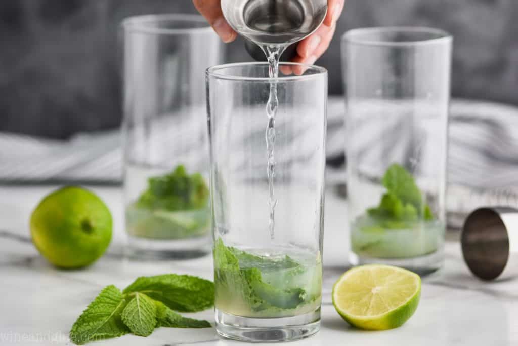 three high ball glasses with muddled mint and lime juice in them to make a mojito recipe and white rum being poured in the first glass