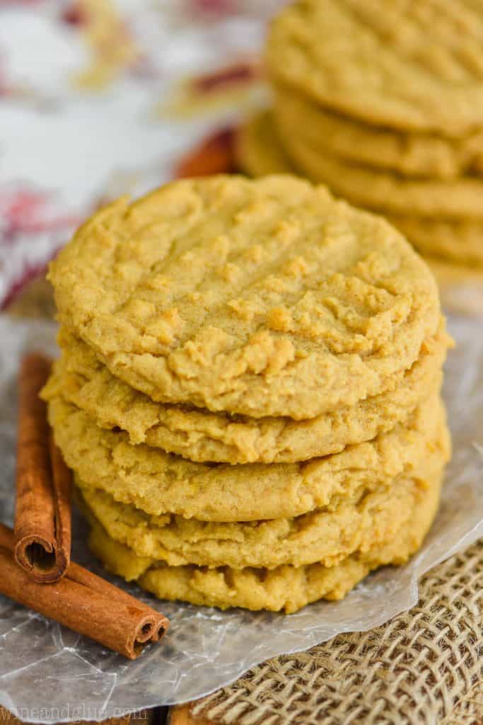 a stack of pumpkin peanut butter cookies on wax paper with burlap underneath, cinnamon sticks next to them, another stack in the background