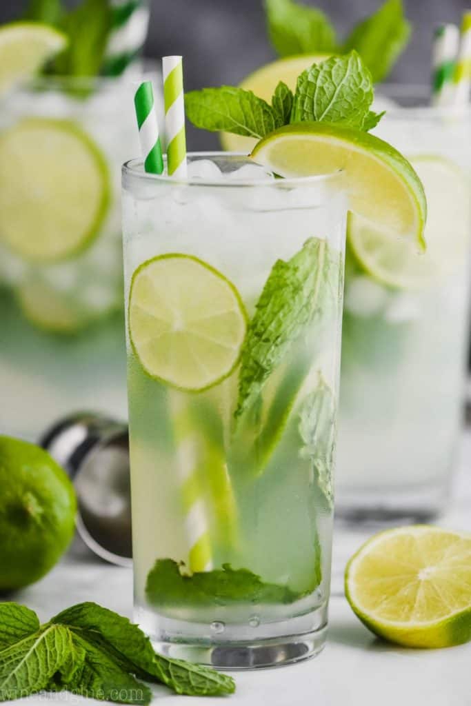 up close view of a high ball glass filled with a mojito cocktail, lime slices, fresh mint, striped green straws, and garnished with lime wedges and fresh mint