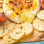 a wheel of baked brie with cheese coming out and topped with marmalade, honey, and pecans on a platter with baguette slices, crackers, and grapes, says "the best baked brie simplejoy.com"