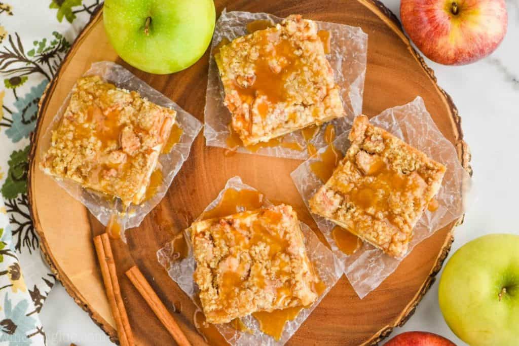 an overhead view of four apple cheesecake bars on wax paper on a circle wood platter