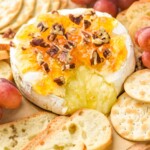 a wheel of baked brie with cheese coming out and topped with marmalade, honey, and pecans on a platter with baguette slices, crackers, and grapes