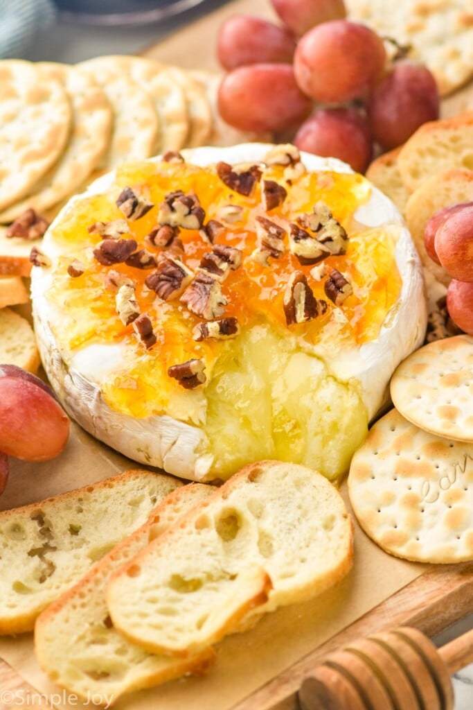 a wheel of baked brie with cheese coming out and topped with marmalade, honey, and pecans on a platter with baguette slices, crackers, and grapes