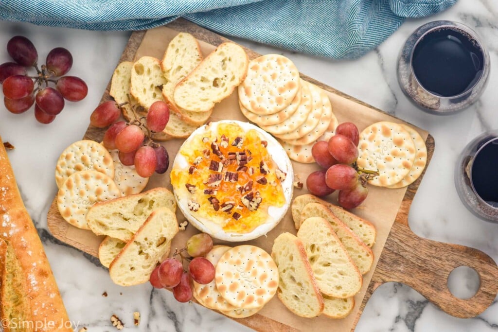 wheel of baked brie on a platter with crackers, baguettes, and grapes, two wine glasses in the photo